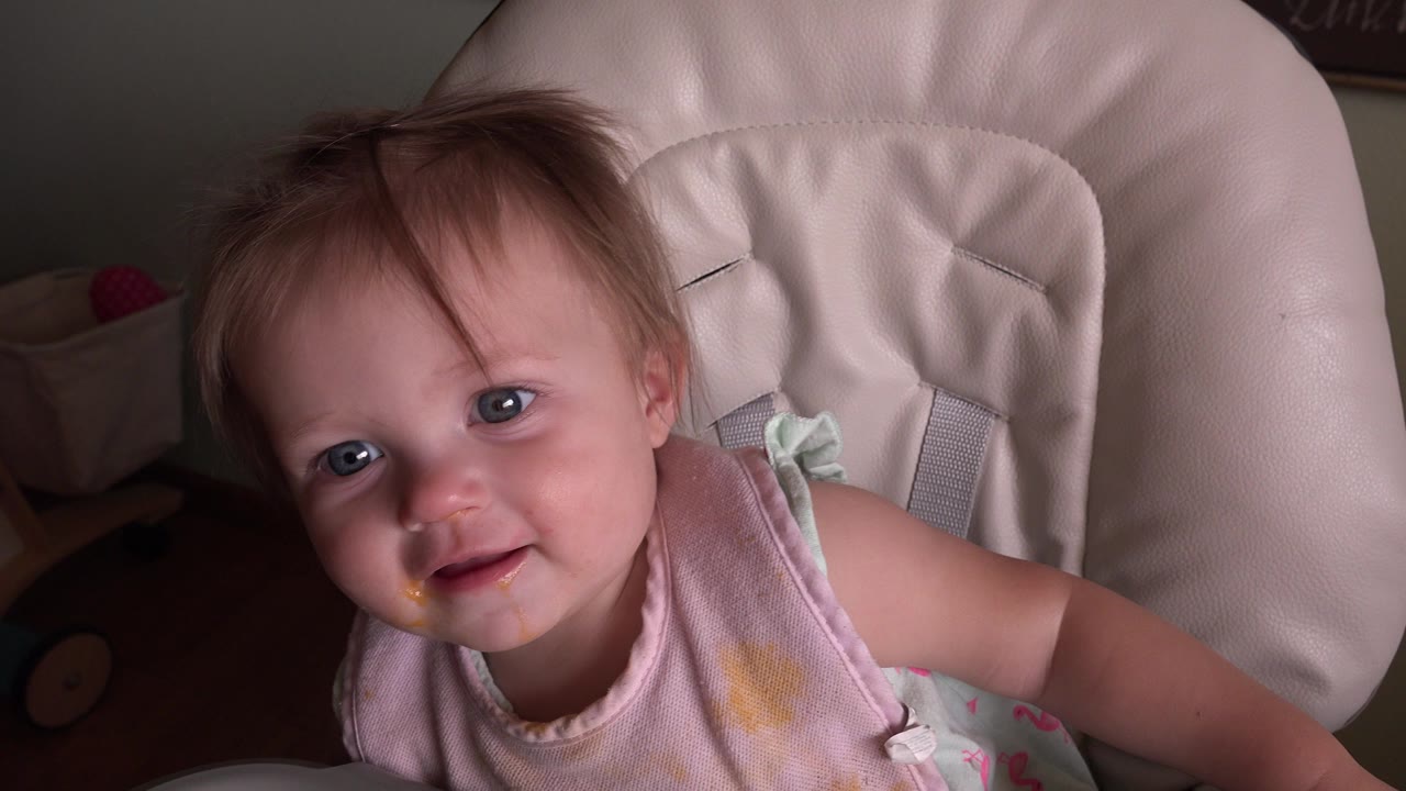 Blond toddler in a pale high chair looking up at the camera.