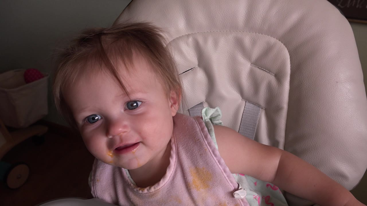 At 0:01, the young blond girl remains close to camera in the high chair, with her head tilted and yellow food marks visible on her mouth and clothing.