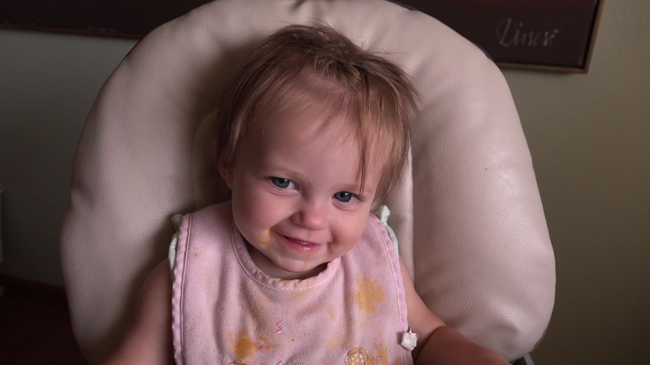 At 0:01, the baby remains centered in the padded highchair, with a slight smile and the pink bib catching the dim indoor light in Hidden Lake, Colorado.