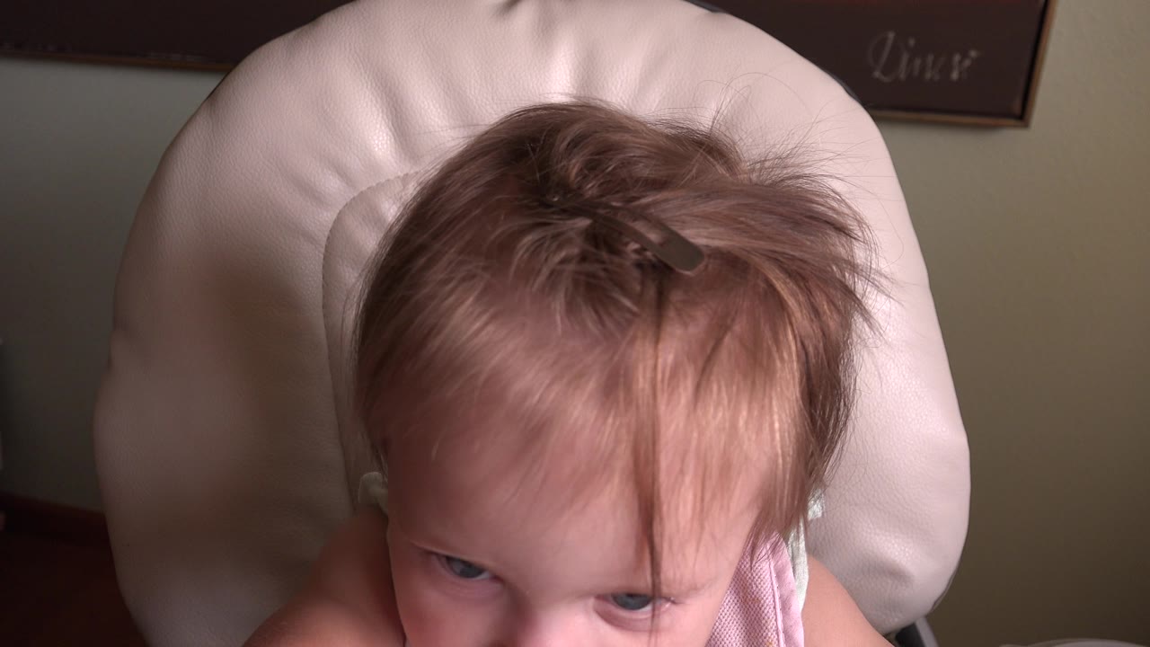At 0:00 in Hidden Lake, Colorado, a toddler peers upward from a pale high chair, with fine hair held by a small clip and muted wall art behind.