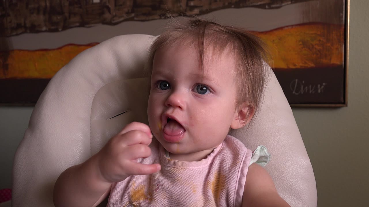 Baby sitting in a cushioned chair with one hand raised near the mouth.