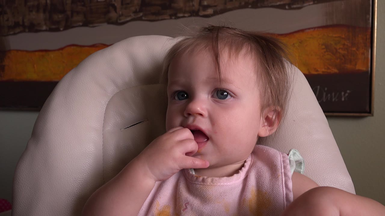 A blond baby sits in a pale high chair at Hidden Lake Colorado, fingers at the mouth, with framed wall artwork behind the chair.