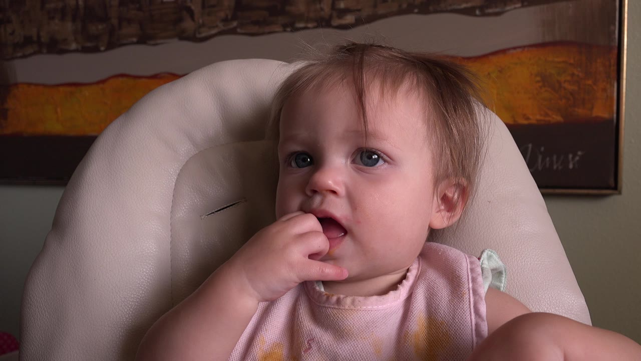 The blond baby remains in the high chair at Hidden Lake Colorado, looking slightly upward with fingers resting at the lips and wall art in the background.