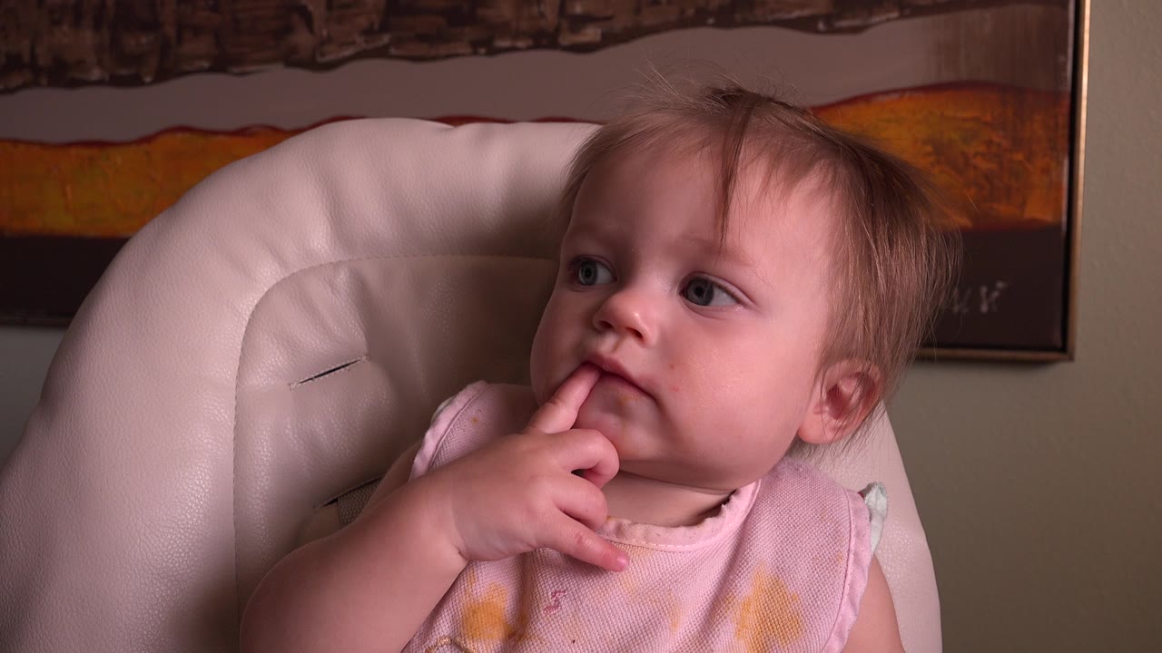 At 0:01, the toddler remains in the high chair in Hidden Lake, Colorado, with one finger in the mouth and eyes turned toward the side of the room.