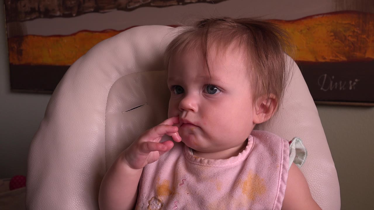 Young child in a pale high chair with fingers near the mouth.
