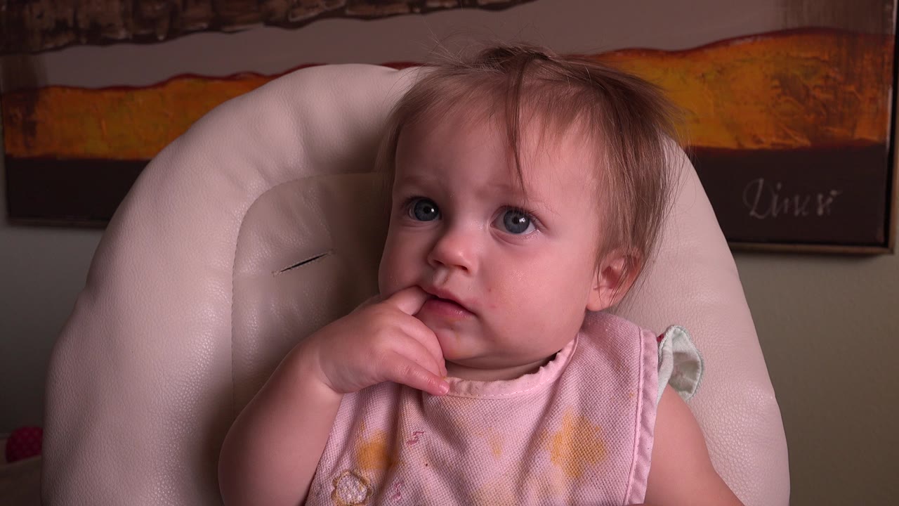 Baby with blue eyes chewing a finger while seated in a pale chair.