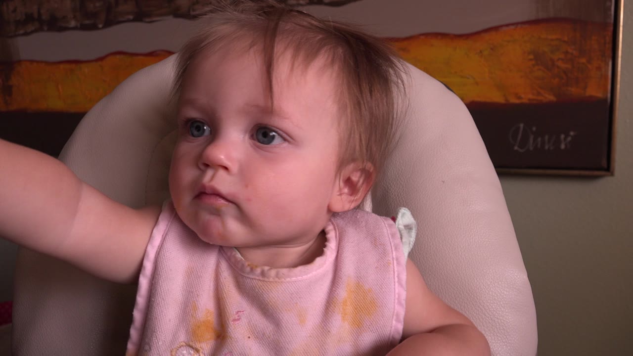 Baby seated in a pale padded chair with one arm reaching left.