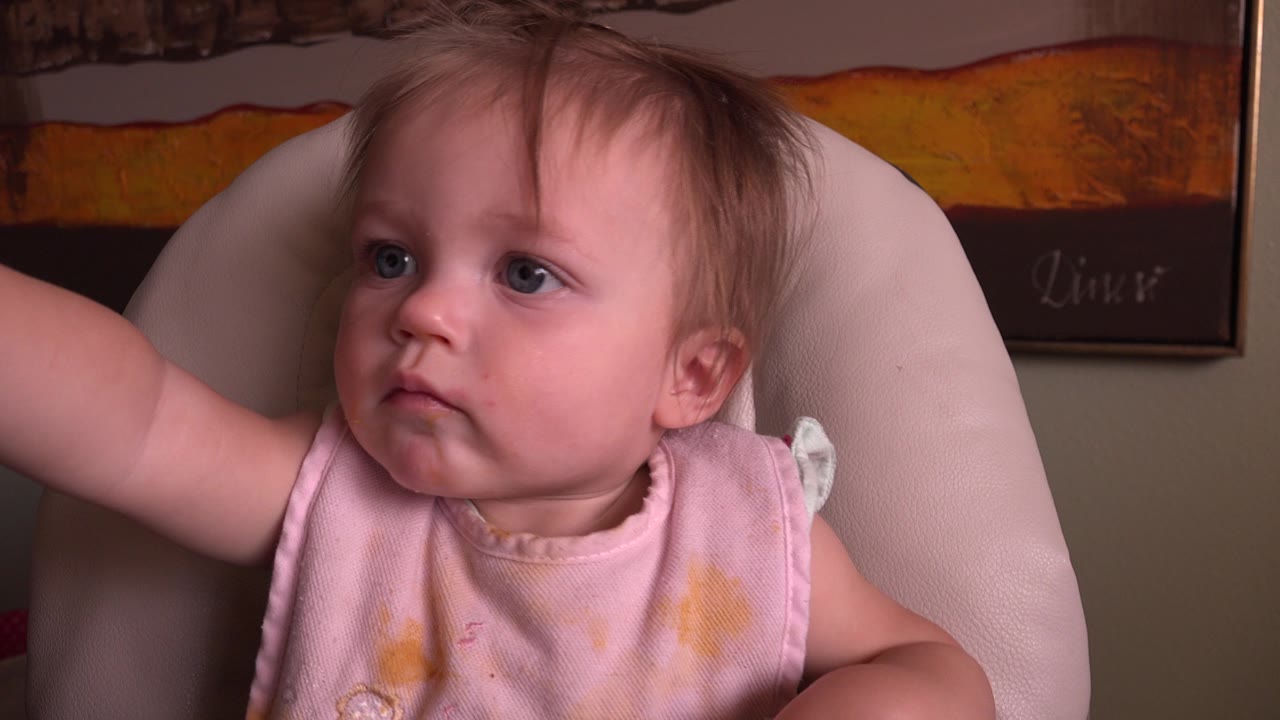 At 0:01, the baby remains in the padded chair, looking left with a raised arm and framed artwork behind the chair.