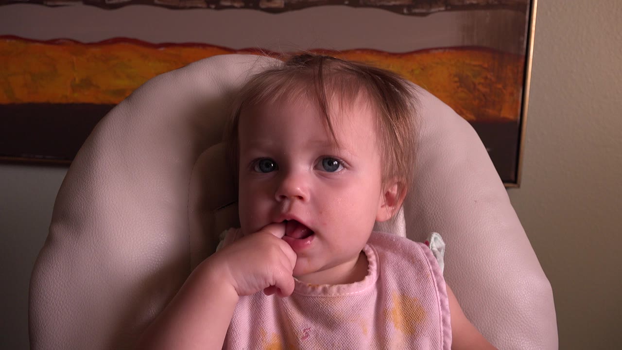 At 0:00, a light-haired baby in Hidden Lake, Colorado sits in a pale padded highchair with fingers near their mouth and orange-brown wall artwork behind them.