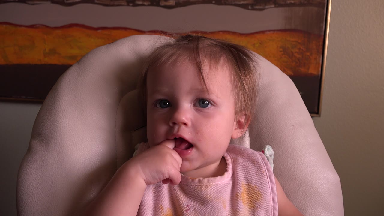 At 0:01, the baby remains centered in the highchair, looking toward the camera with one hand at their mouth beneath the framed artwork.