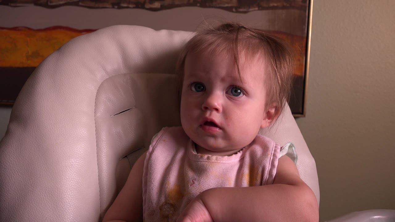 At 0:01, the young child remains centered in the pale chair, eyes lifted and mouth slightly open against the beige interior wall.