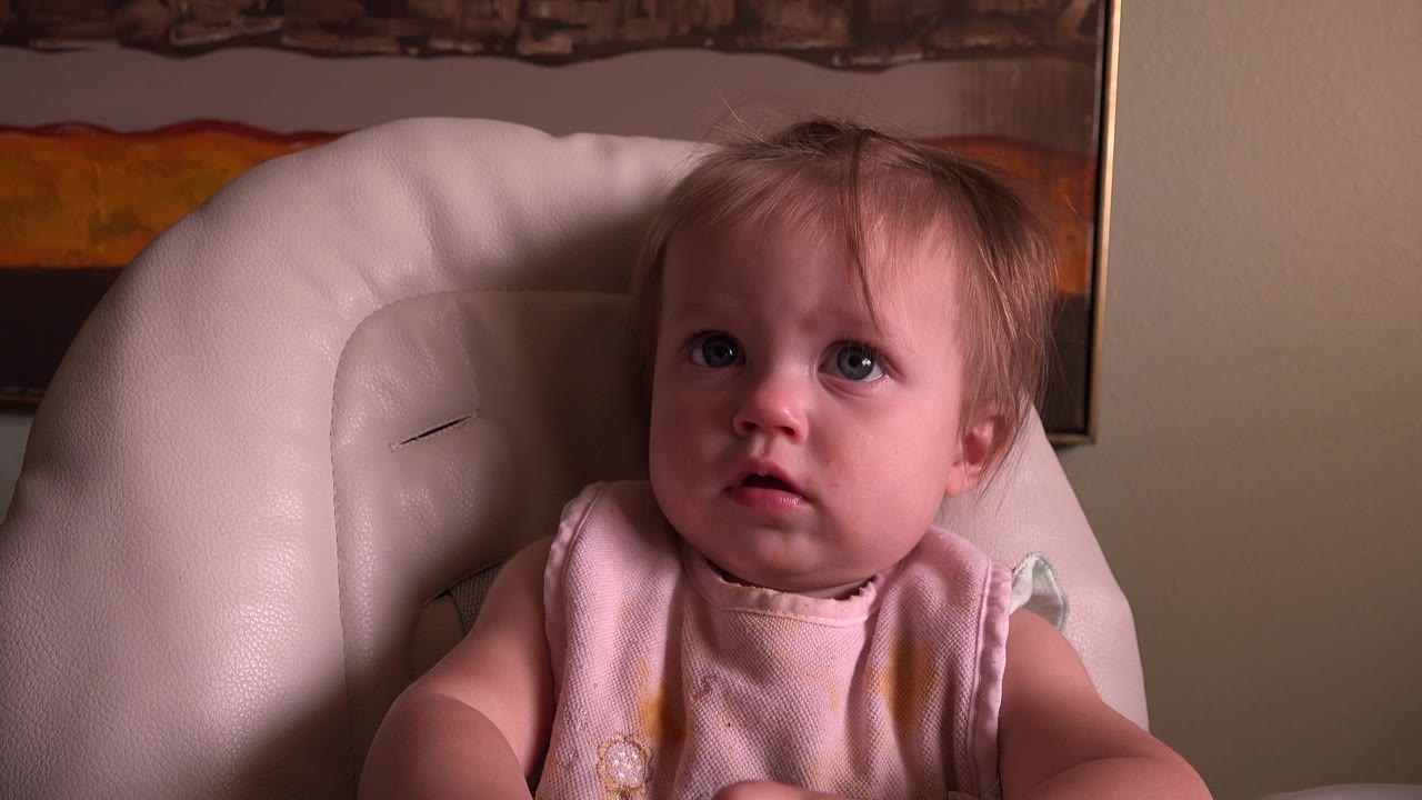 A blond baby with blue eyes sits in a pale pink cushioned chair in Hidden Lake, Colorado, looking upward beneath framed wall art.