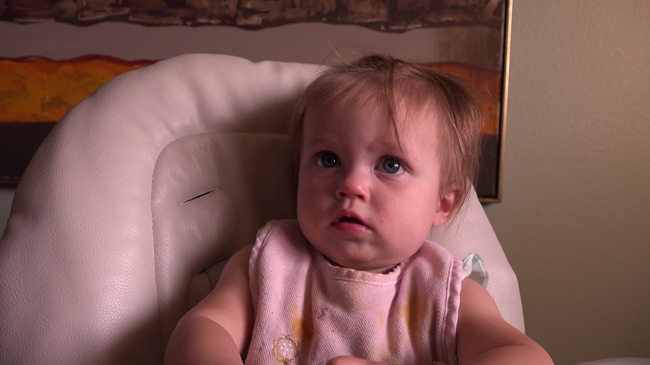 The baby remains seated in the pink chair in Hidden Lake, Colorado, with blue eyes turned upward and warm indoor light on the face.