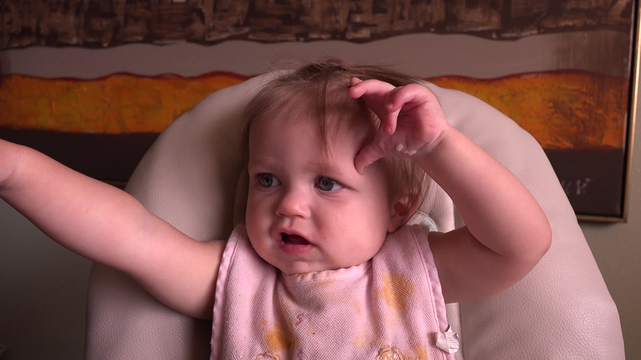 Baby in a chair with one arm stretched outward and the other hand near the head.