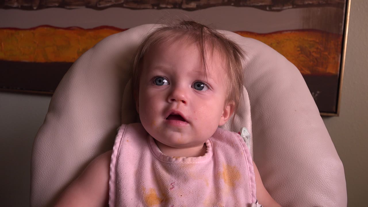 At 0:00, a baby girl in a pale pink outfit sits in a light cushioned chair at Hidden Lake, Colorado, looking upward with a framed artwork behind her.