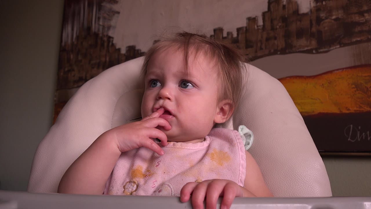 The baby remains seated in the high chair in Hidden Lake, Colorado, looking up with fingers touching her lips and artwork on the wall behind.