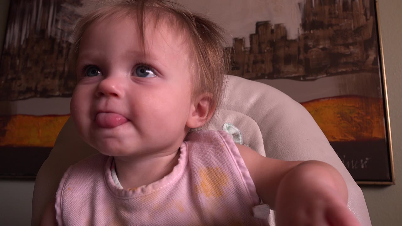At 0:01, the baby remains seated in the pale chair with tongue out, eyes turned upward, and one arm near the right edge of the frame.