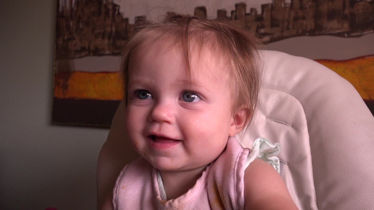 A baby in a pale pink bib smiles from a white high chair inside a Hidden Lake, Colorado home setting, with framed artwork visible behind.