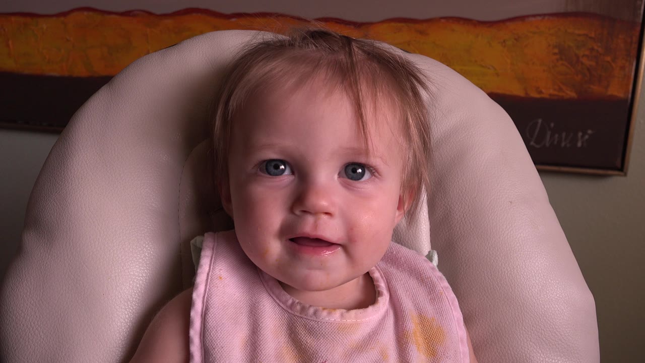 Baby girl in a pink bib sitting in a cushioned chair.