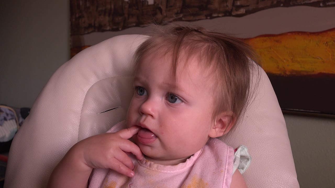 At 0:01, the toddler remains seated in the padded chair in the Hidden Lake, Colorado home movie, looking off frame while holding fingers against the lips.