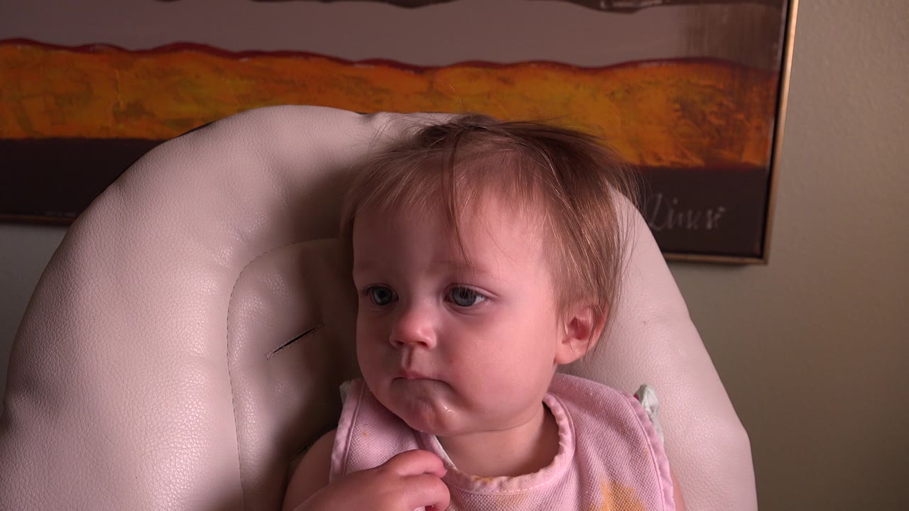 At 0:00, a young girl in a pink bib sits low in a pale padded armchair in a Hidden Lake, Colorado home interior, with warm framed artwork on the wall behind her.