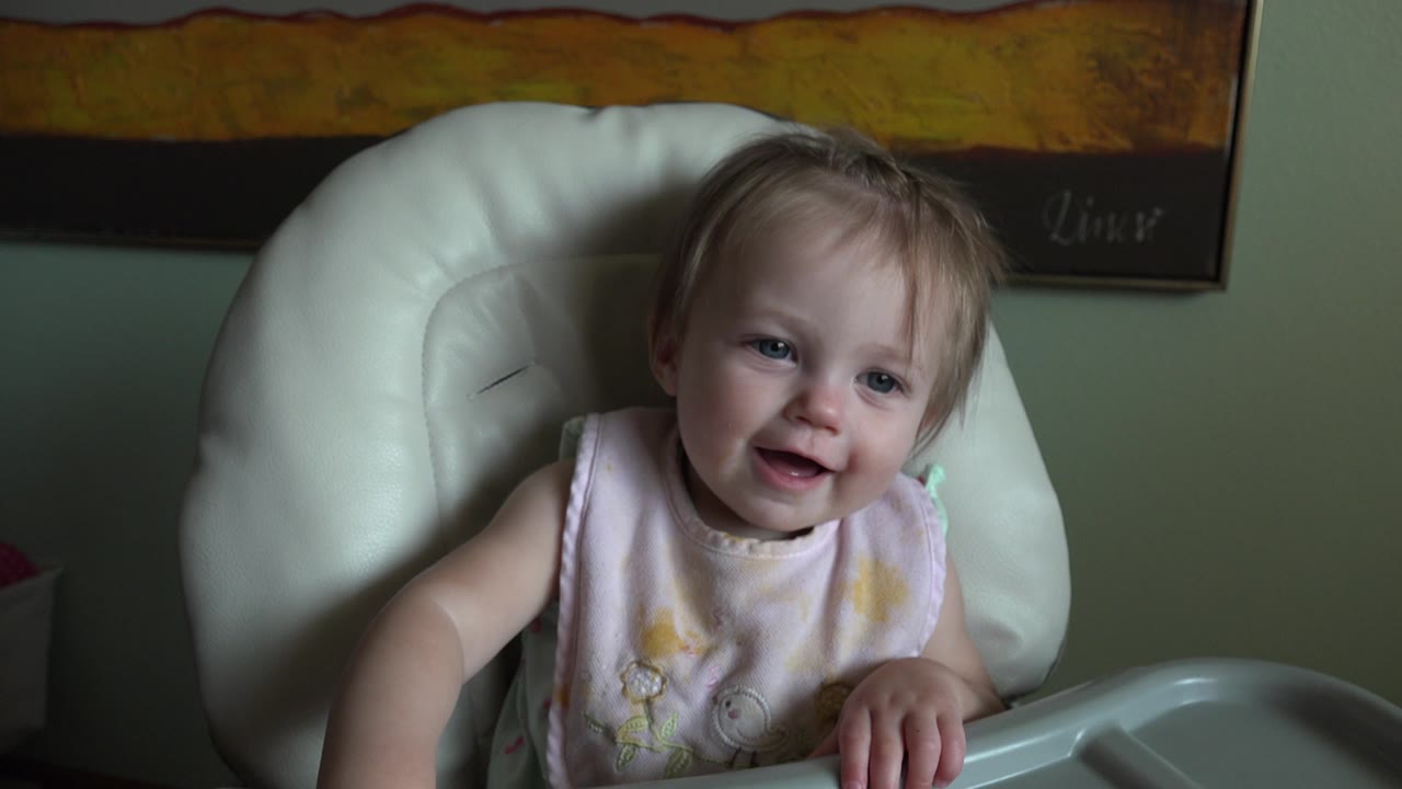 The child remains centered in the white high chair, smiling with one hand resting near the tray in the dim indoor room in Hidden Lake, Colorado.