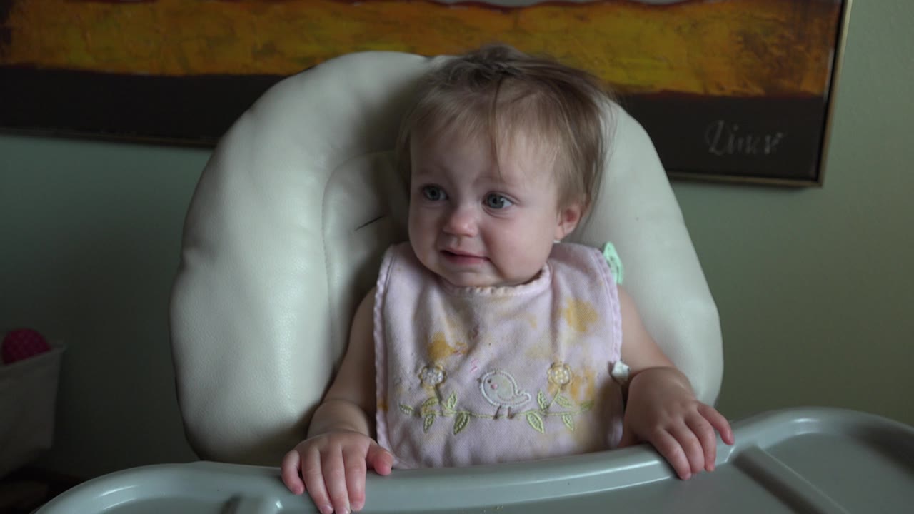 At 0:00, a young girl in Hidden Lake, Colorado sits in a white padded high chair and looks left with her hands on the tray.