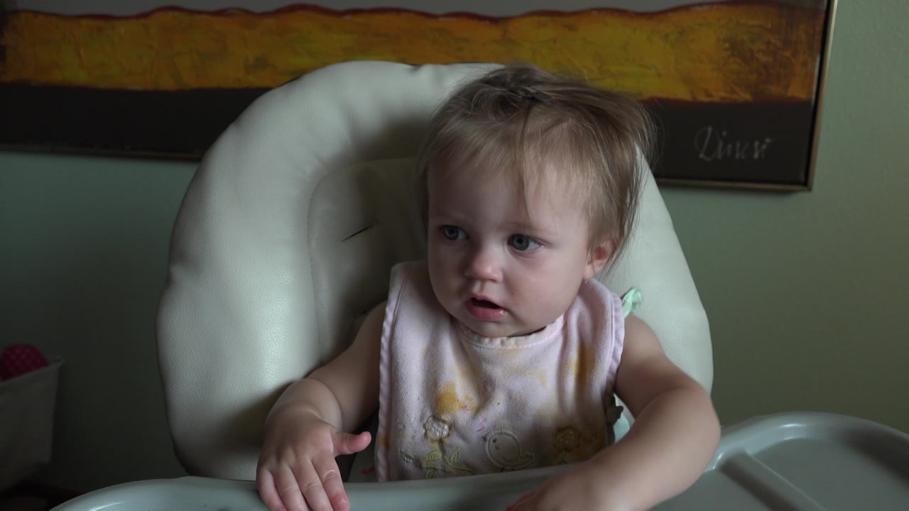 At Hidden Lake, Colorado in 2015, the blond baby remains in the high chair, looking off left while one hand lifts near the tray and framed wall art sits behind.