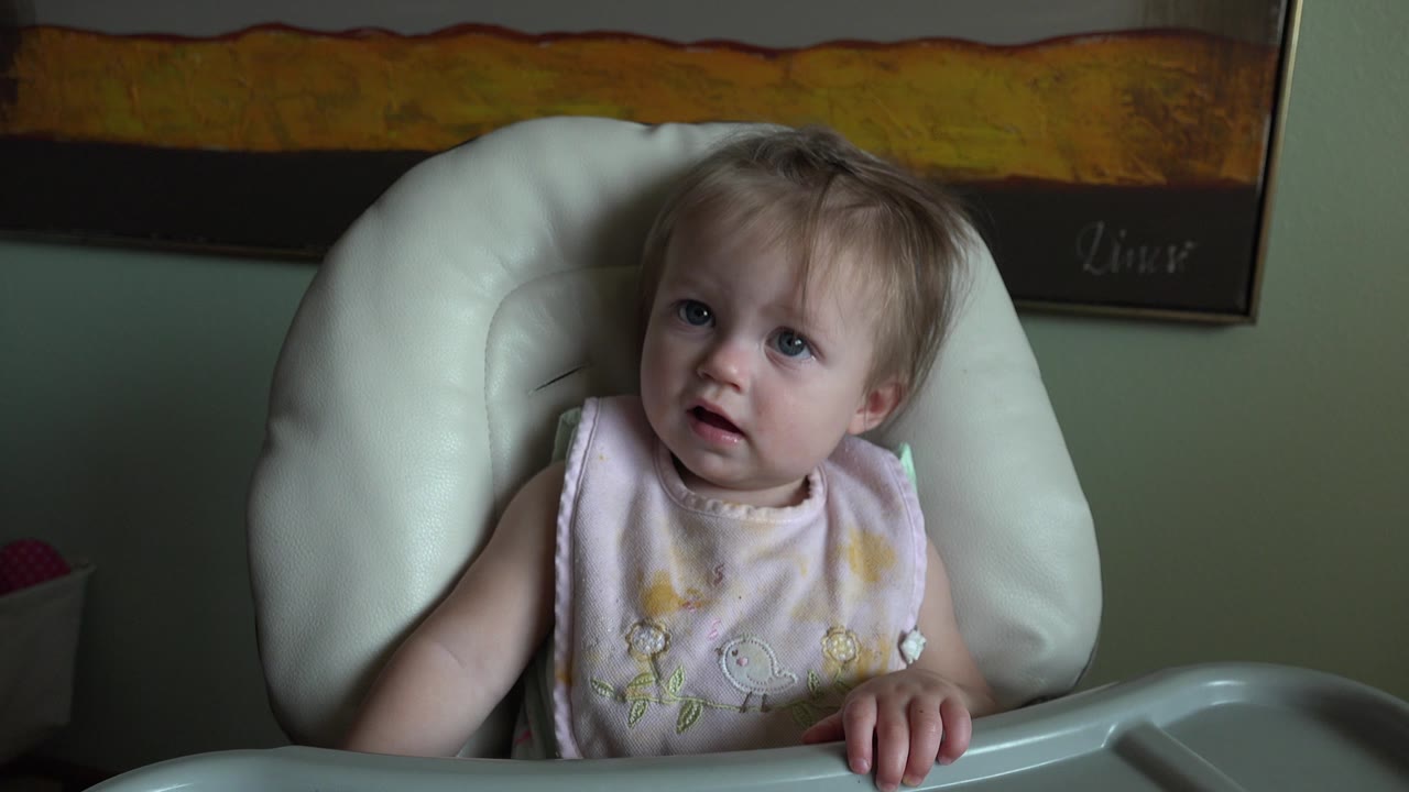 At 0:01, the child looks slightly upward while seated in the white high chair, with soft indoor light on the face and wall art above.