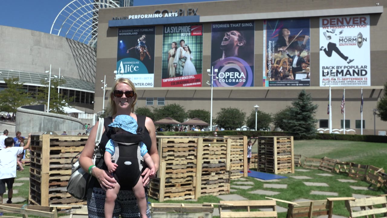 At 0:00 in Denver, Colorado, a woman in sunglasses stands with a baby in a black front carrier, with wood pallets and large performing arts posters behind her.