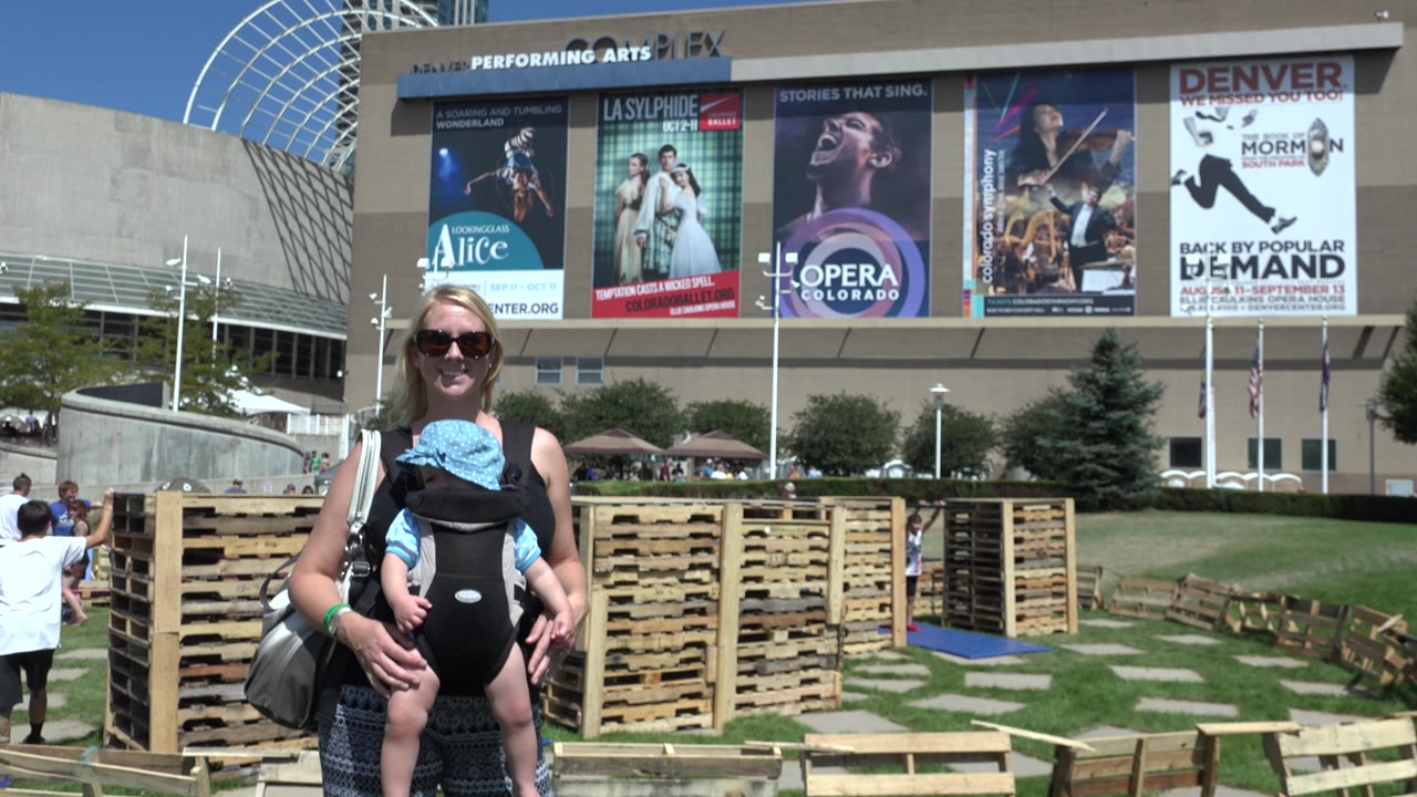 At 0:01 in Denver, Colorado, the woman and baby remain centered, with the baby facing forward in the carrier and the venue banners visible across the building facade.