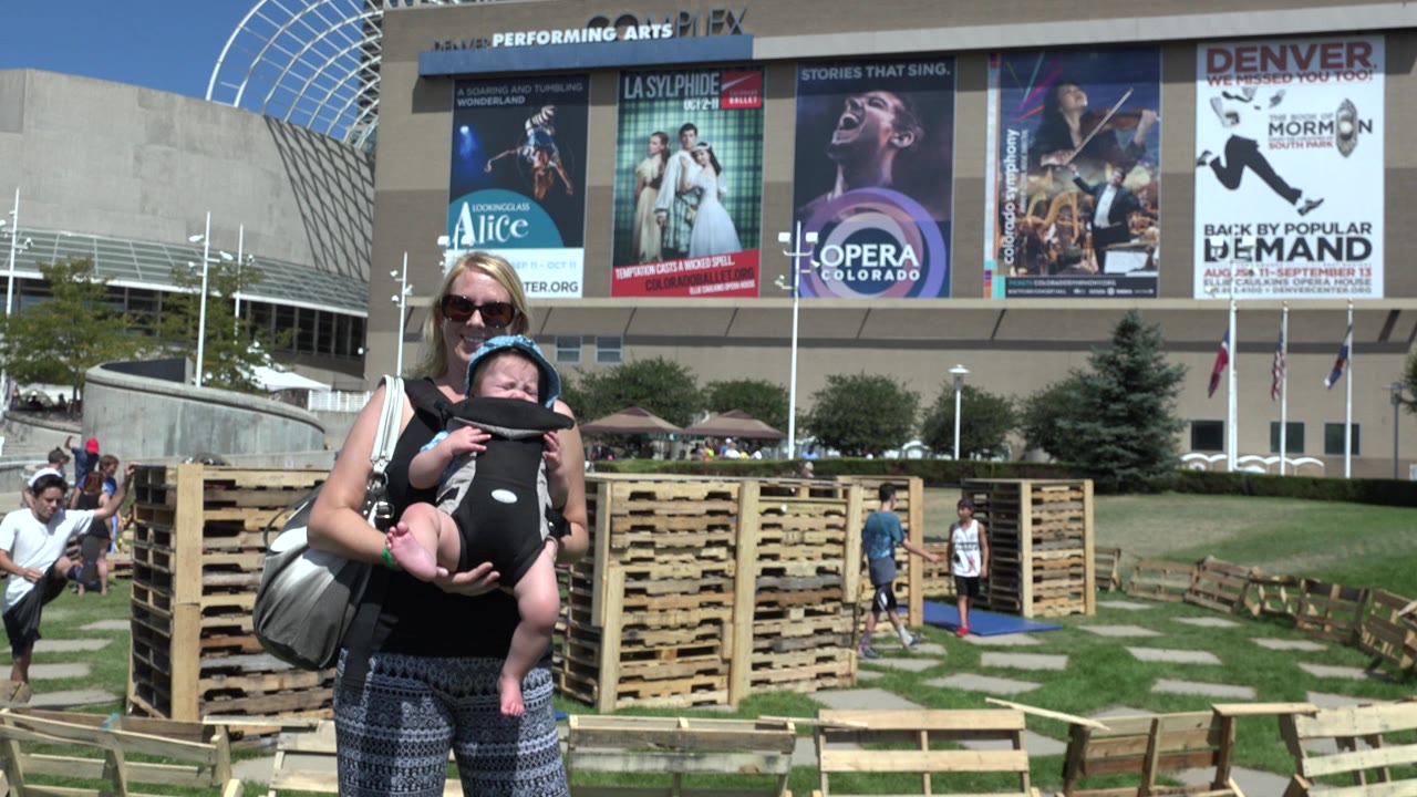 Woman in sunglasses holds a baby in a front carrier outside a large arts venue.