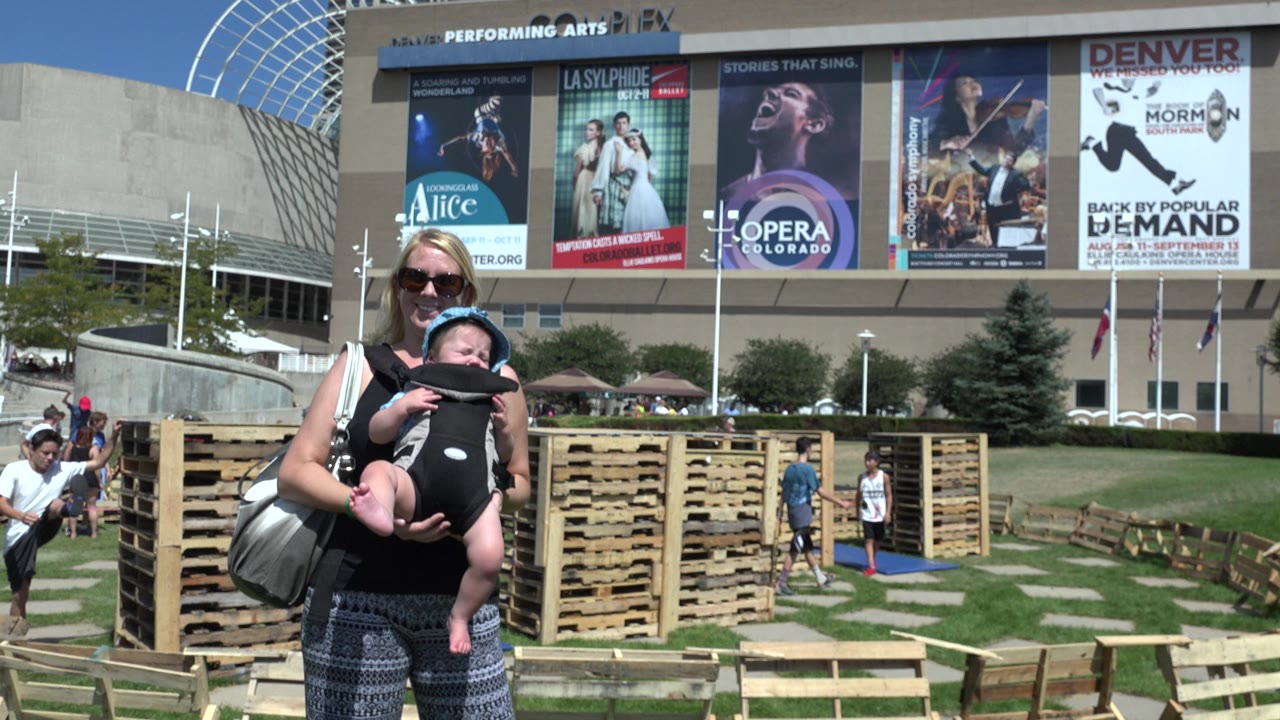 In Denver, the woman and baby remain centered while large performance posters, a lawn, wood pallet structures, and a few people appear in the background.
