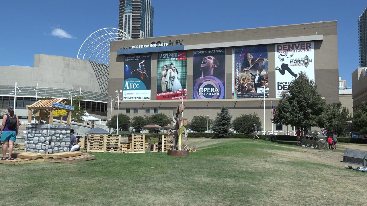 Wide grassy plaza outside a performing arts building with posters and scattered people.