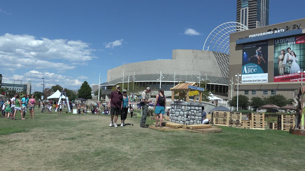 Wide view of a Denver outdoor exhibition field with attendees walking near a stone-well style display, white tents, and a large venue building under bright sun.