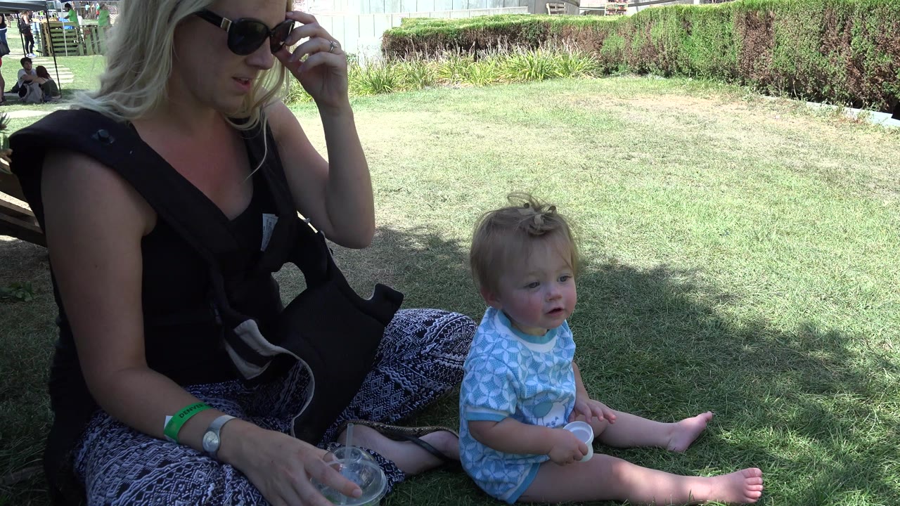 In Denver, Colorado, a blonde woman wearing sunglasses sits on shaded grass beside a toddler in a light blue outfit, with a bright lawn and hedges behind them.