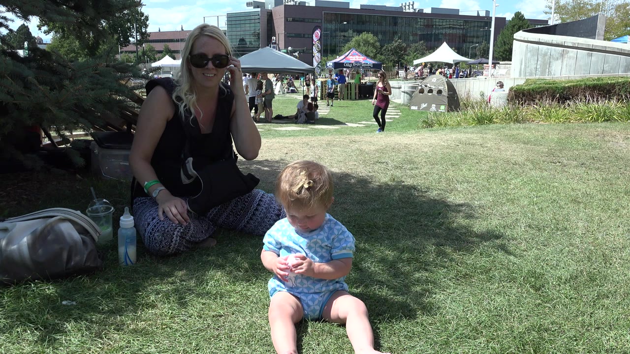 In Denver, a blonde woman in sunglasses sits in shade behind a toddler on grass while tents, pedestrians, concrete features, and buildings fill the sunlit background.