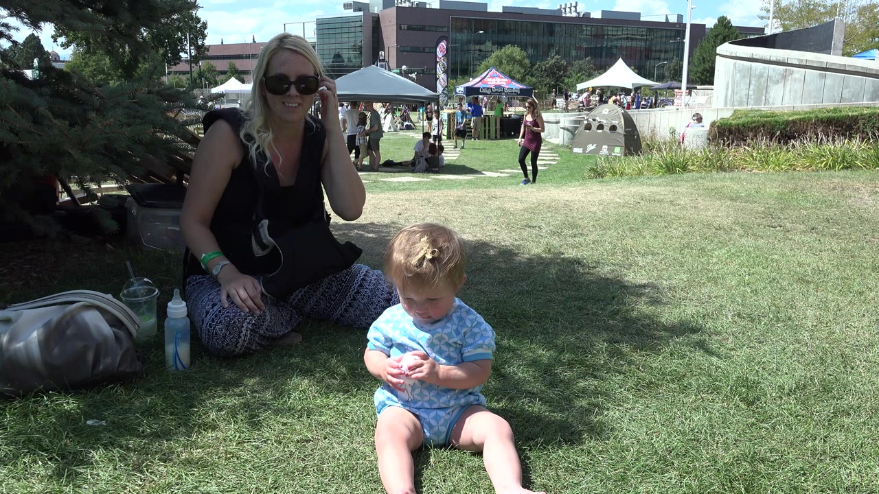 In Denver, the seated woman smiles from the shaded left side of the frame as the toddler sits in the foreground looking down, with tents and people in bright sun behind them.