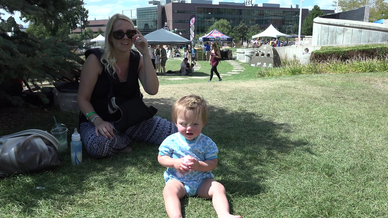 In Denver, the toddler sits more upright near the lower right foreground while the mother remains seated in shade, with a lawn, tents, walking people, and office-like buildings behind them.