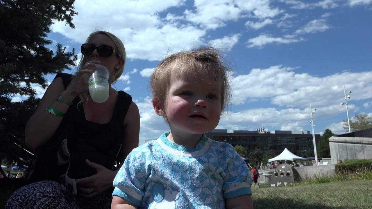 Toddler seated on grass beside a woman drinking from a bottle.