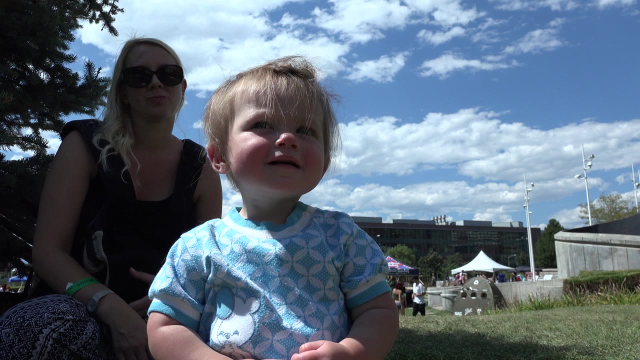 Infant sitting on grass with woman behind her under a partly cloudy sky.
