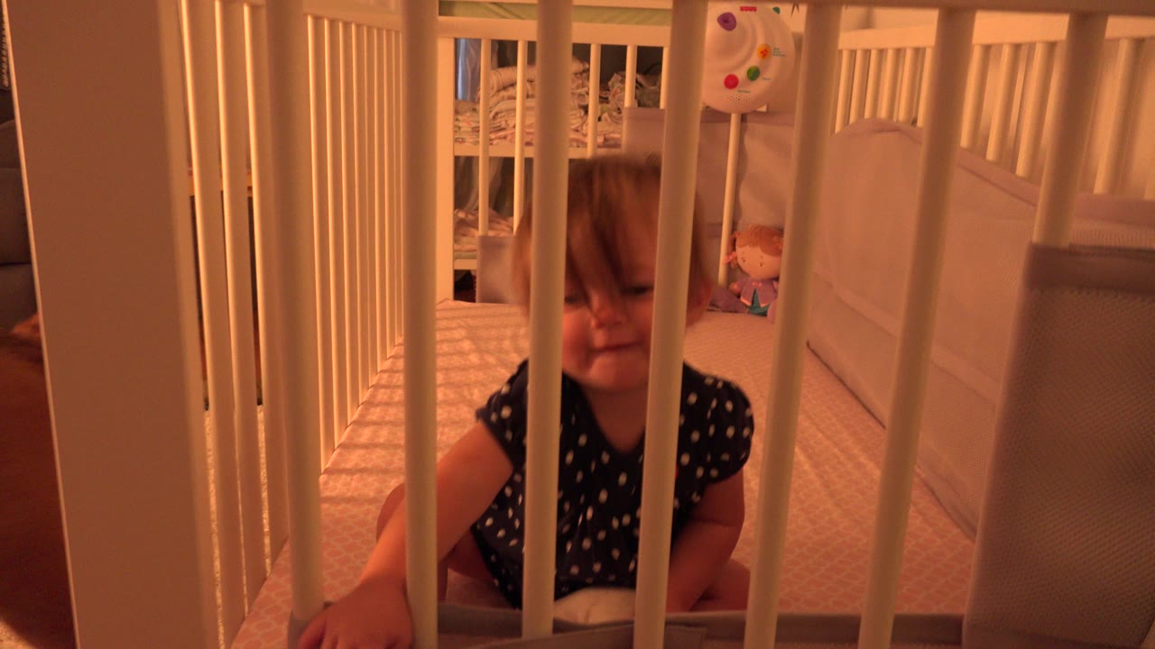 The toddler remains seated behind the white crib rails in the Westminster, Colorado home interior, with warm orange light and bedding visible behind the bars.