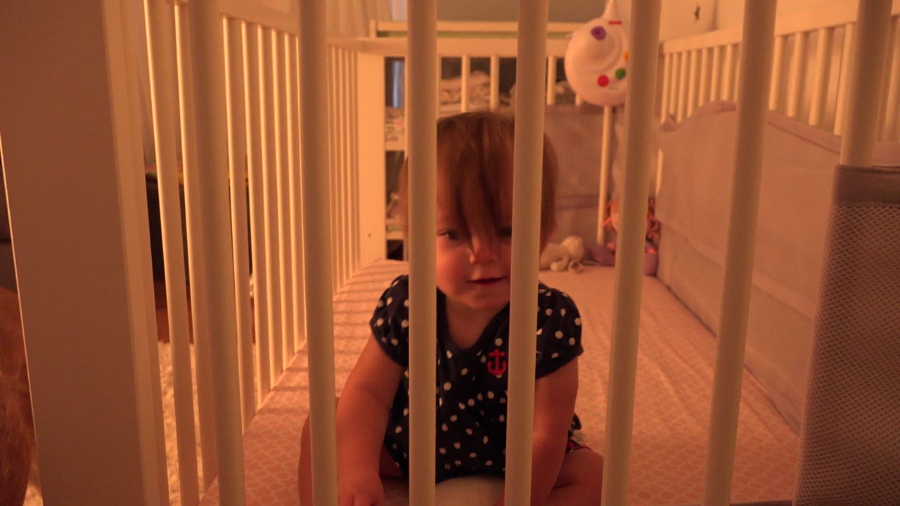 Toddler girl sits behind white crib rails inside a Westminster, Colorado home, with beige carpet and a soft toy visible in the warm-lit background.