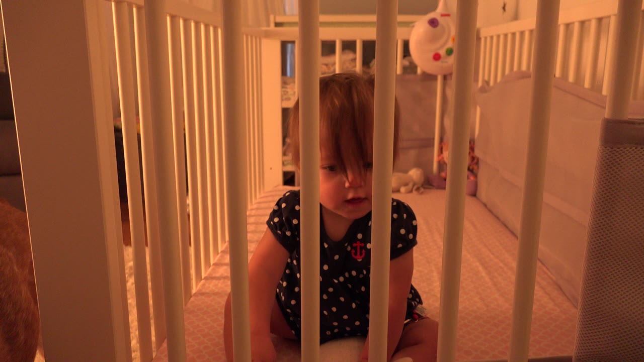 Toddler girl behind crib rails with a white crib side in the foreground.