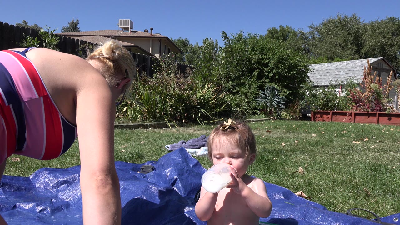 Woman leaning over a toddler drinking from a bottle on a blue tarp.
