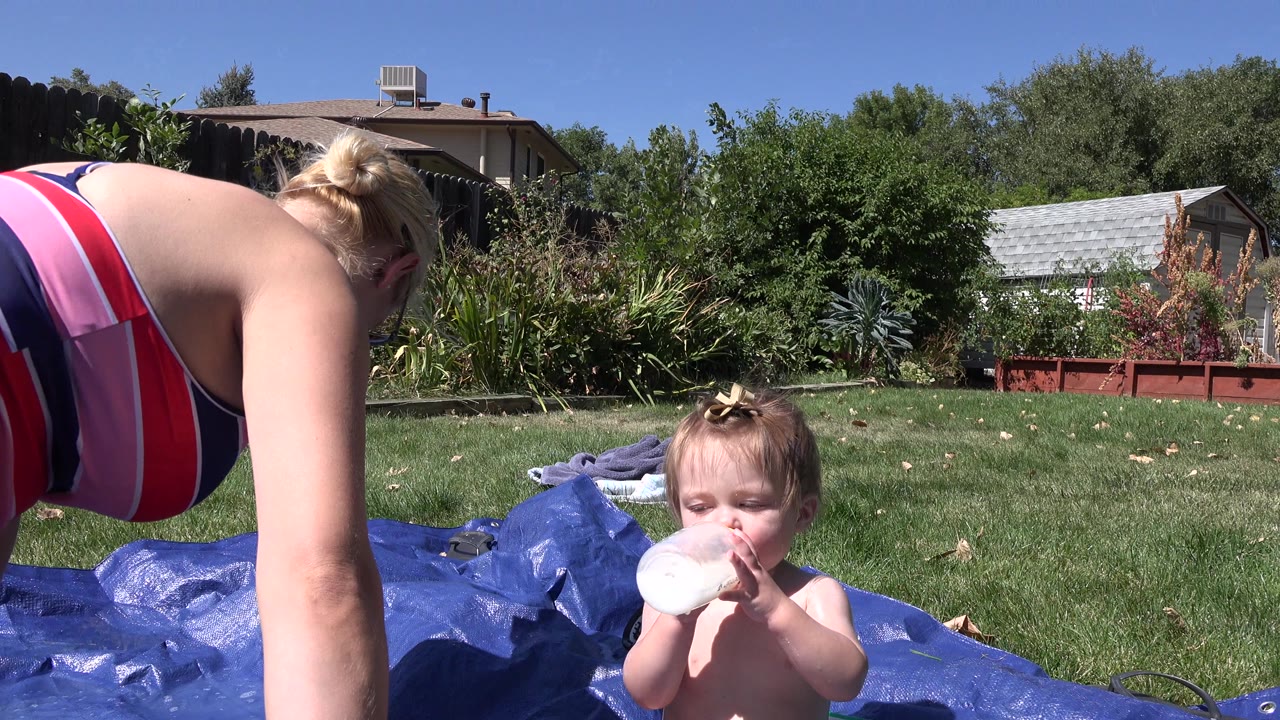 The toddler remains centered on the blue tarp drinking from a bottle while the woman bends close at the left edge of the sunny Westminster backyard frame.