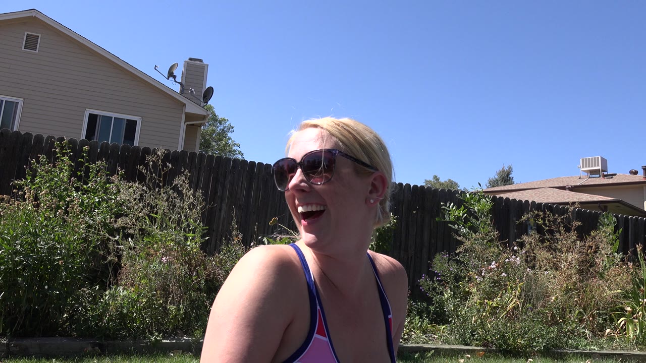 A blonde woman in sunglasses smiles near the camera in a Westminster, Colorado backyard, with a wooden fence, garden plants, and neighboring houses behind her.