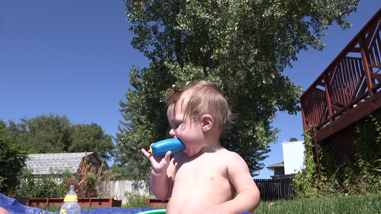 Toddler holds a blue toy near their mouth on a backyard water mat.