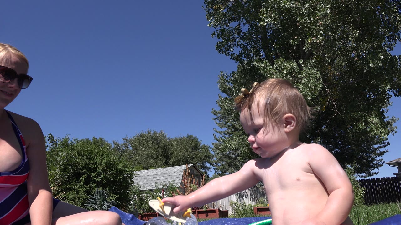 A baby reaches toward toys or water items on a blue mat while his mother sits partly in frame in a Westminster, Colorado backyard under bright sun.