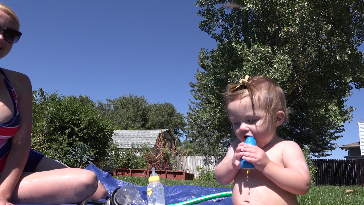 At 0:01 in Westminster, Colorado, the baby faces forward and bites the blue treat as the adult remains partly cropped at the left and yard objects sit along the pool edge.
